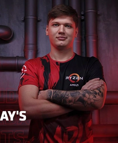 Een esports-speler in een rood-zwart tenue staat met zijn armen over elkaar tegen de achtergrond van een industrieel interieur